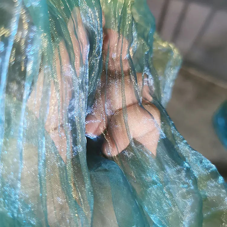 Une main appuie sur le tissu semi-transparent et brillant bleu-vert Tissu organza irisé plissé au mètre de Longan Craft France, laissant apparaître les doigts et les articulations à travers le délicat tissu dégradé bicolore.