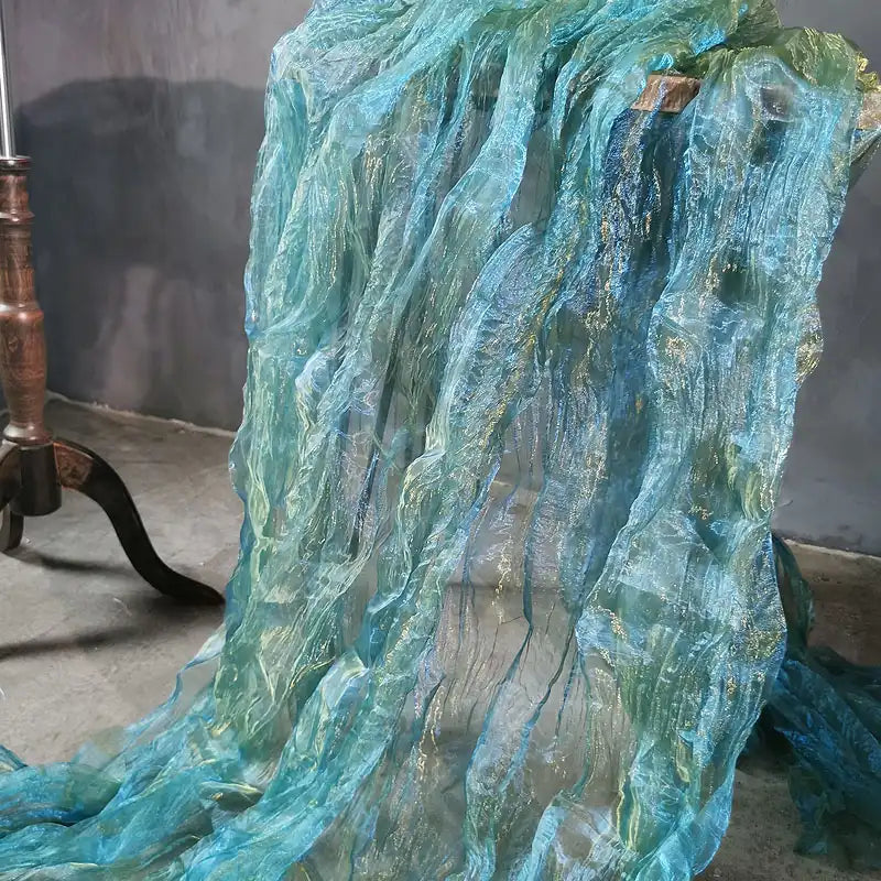 Le tissu Longan Craft France Tissu organza irisé plissé au mètre, un tissu bicolore transparent avec un motif ondulé chatoyant bleu et vert, se drape sur des meubles à pieds en bois et s'étale sur un sol en béton gris.