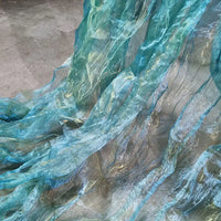 Gros plan sur le Tissu organza irisé plissé au mètre de Longan Craft France, un tissu dégradé transparent bicolore bleu et aqua, drapé sur du gris pour créer un effet d'ondulation et d'eau.