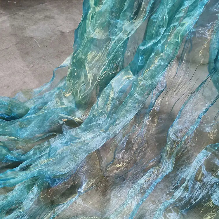Gros plan sur le Tissu organza irisé plissé au mètre de Longan Craft France, un tissu dégradé transparent bicolore bleu et aqua, drapé sur du gris pour créer un effet d'ondulation et d'eau.