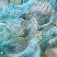 Gros plan sur un tissu bleu-vert translucide et texturé, dont les couches ondulées évoquent l'eau qui coule, comme le Tissu organza irisé plissé au mètre de Longan Craft France, un tissu bicolore transparent à l'effet de dégradé chatoyant.