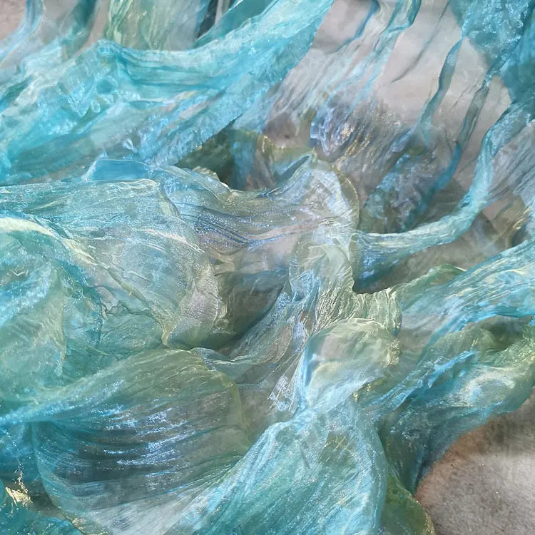 Gros plan sur un tissu bleu-vert translucide et texturé, dont les couches ondulées évoquent l'eau qui coule, comme le Tissu organza irisé plissé au mètre de Longan Craft France, un tissu bicolore transparent à l'effet de dégradé chatoyant.