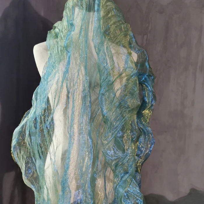 Un mannequin blanc présente le Tissu organza irisé plissé au mètre de Longan Craft France, un tissu bicolore transparent aux dégradés de bleu et de vert qui scintille joliment sur un fond sombre.