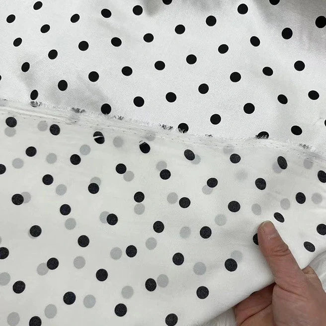 Gros plan sur le tissu en soie extensible à pois au mètre de Longan Craft France, une soie imprimée avec élasthanne d'inspiration vintage, en blanc et noir, partiellement voilée par une couche transparente à pois alors qu'une main en soulève délicatement le bord.