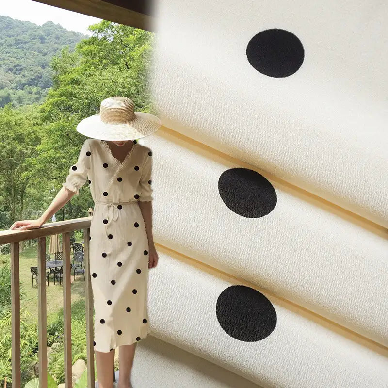 Une personne vêtue d'une robe blanche en "Tissu viscose fleuri à pois" de Longan Craft France et d'un chapeau de paille à larges bords se tient sur un balcon. Un gros plan du tissu 100 % viscose est montré à droite, avec des arbres verts en arrière-plan.