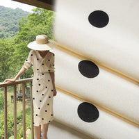 Une personne vêtue d'une robe blanche en "Tissu viscose fleuri à pois" de Longan Craft France et d'un chapeau de paille à larges bords se tient sur un balcon. Un gros plan du tissu 100 % viscose est montré à droite, avec des arbres verts en arrière-plan.