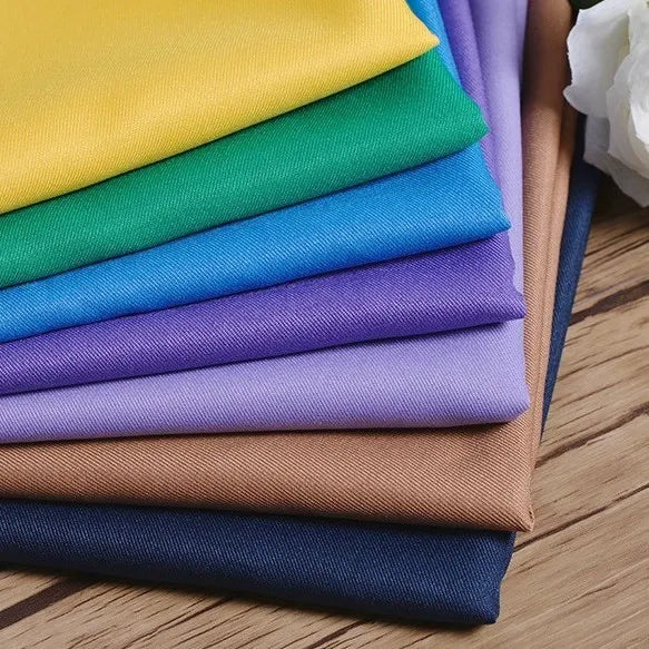 Une pile de Tissu gabardine 100 % polyester au mètre de Longan Craft France en jaune, vert, bleu, violet, beige et marine - idéal pour les vestes élégantes - repose sur une surface en bois avec une fleur blanche partiellement visible.