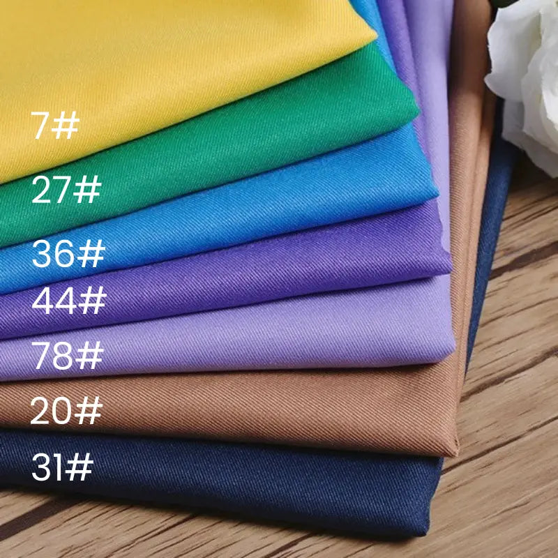 Une pile de tissu gabardine 100% polyester au mètre de Longan Craft France, dans les coloris 7#, 27#, 36#, 44#, 78#, 20# et 31#, bien rangés sur une surface en bois à côté d'une fleur blanche - idéal pour les vestes élégantes.
