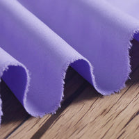 Gros plan sur le Tissu gabardine 100% polyester au mètre de Longan Craft France, de couleur violet clair. Le tissu sergé uni de qualité est drapé en vagues douces sur le bois, montrant une texture lisse avec une brillance subtile - idéal pour les vestes élégantes.