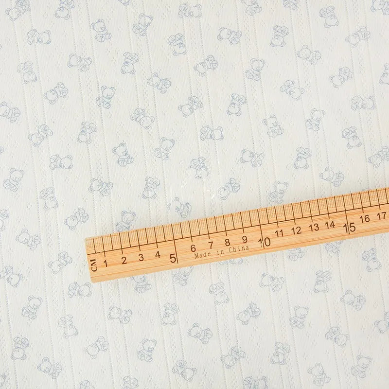 Une règle en bois est posée sur le "Tissu pointelle motif ours au mètre" de Longan Craft France, un tissu de coton léger avec un motif d'ourson ajouré, montrant des marques de centimètres et de pouces. Règle étiquetée "Made in China".