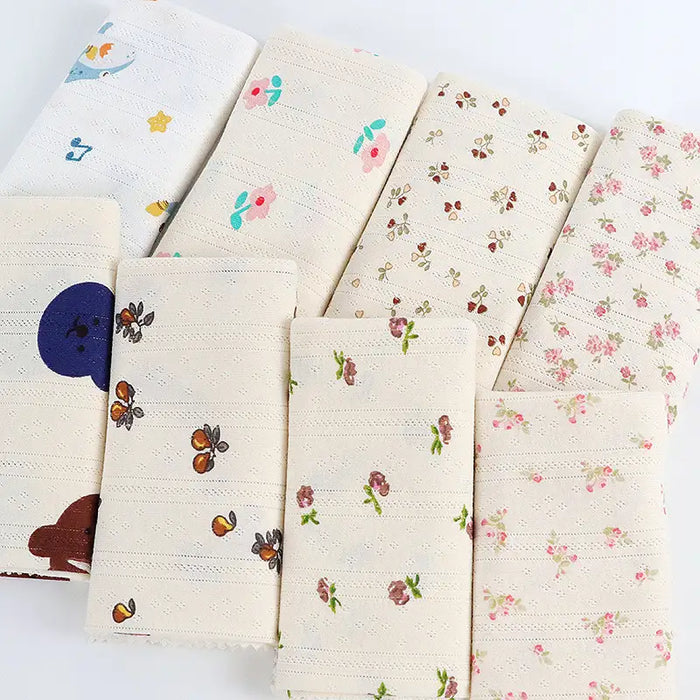 Huit morceaux beiges pliés de Tissu pointelle en coton imprimé au mètre par Longan Craft France, présentant de petits motifs floraux, de papillons et d'animaux en rose, bleu, jaune, marron et vert, sont disposés sur une surface blanche.