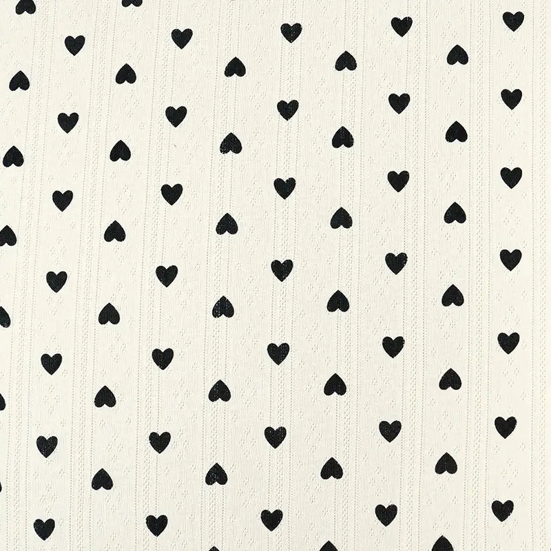 Un fond blanc avec des rangées verticales régulièrement espacées de petits cœurs noirs unis - idéal pour la couture enfant. Tissu pointelle en coton imprimé au mètre de Longan Craft France, maille haute qualité pour vêtements bébé DIY.