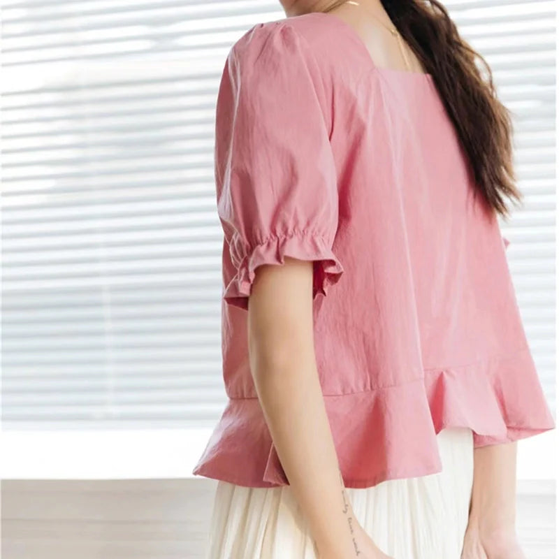 Une femme aux longs cheveux bruns porte le "Patron de couture gratuit pour blouse à manches bouffantes" de Longan Craft France - une blouse rose, ample, à col carré et ourlet à volants - associée à une jupe plissée blanche près d'une fenêtre aux stores blancs.