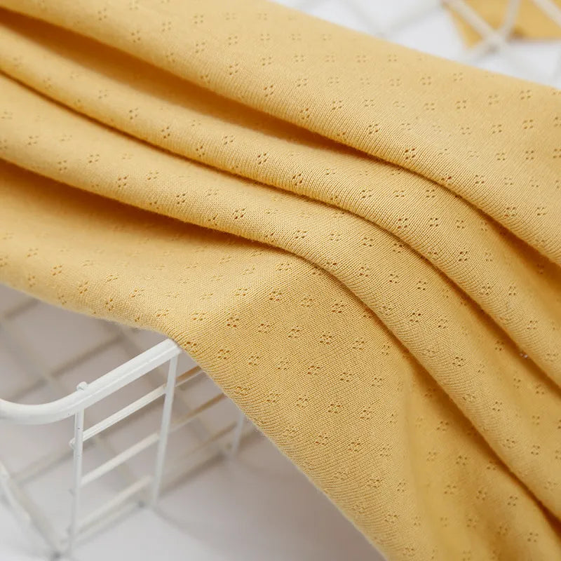 Découvrez le Tissu jersey pointelle 100% coton de Longan Craft France - un tissu jacquard jaune pâle et doux au motif subtil en pointillés, parfait pour les bodys de bébé DIY et drapé ici sur un panier en fil de fer blanc.