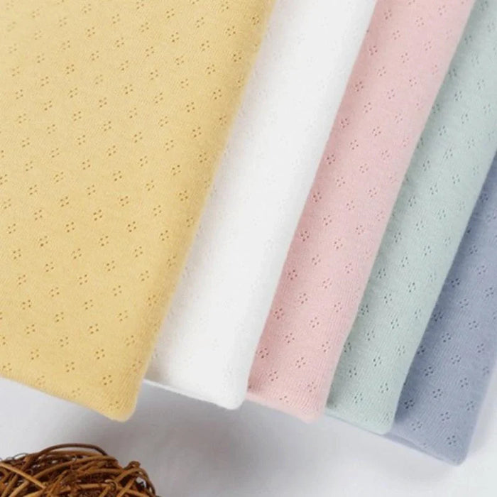 Cinq pièces pliées de Tissu jersey pointelle 100 % coton de Longan Craft France, en jaune, blanc, rose, bleu clair et gris avec un délicat motif d'œillets, sont posées côte à côte ; un panier tressé apparaît en bas à gauche.