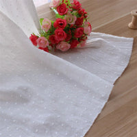 Un bouquet de roses artificielles roses et rouges repose sur une surface en bois à côté d'un mètre de Tissu plumetis 100 % coton blanc pur de Longan Craft France, idéal pour les vêtements légers et la décoration. Une petite bobine apparaît dans le coin supérieur droit.
