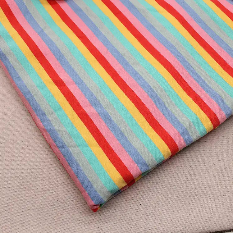 Un morceau de "Tissu French Terry extensible à rayures arc-en-ciel" de Longan Craft France, présentant des rayures rouges, roses, jaunes, bleues et vertes, repose sur une surface beige unie.