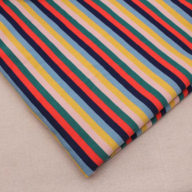 Un morceau plié de Longan Craft France Tissu French Terry extensible à rayures arc-en-ciel, un tissu de coton doux à rayures verticales colorées, est présenté sur une surface beige.