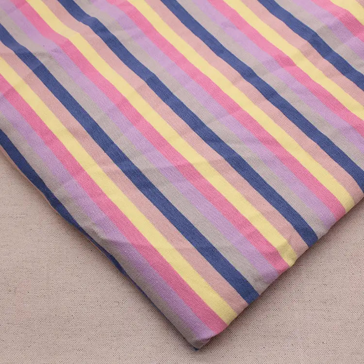 Un morceau de Longan Craft France Tissu French Terry extensible à rayures arc-en-ciel, présentant des rayures verticales bleues, roses, jaunes et lavande, repose sur une surface beige unie.