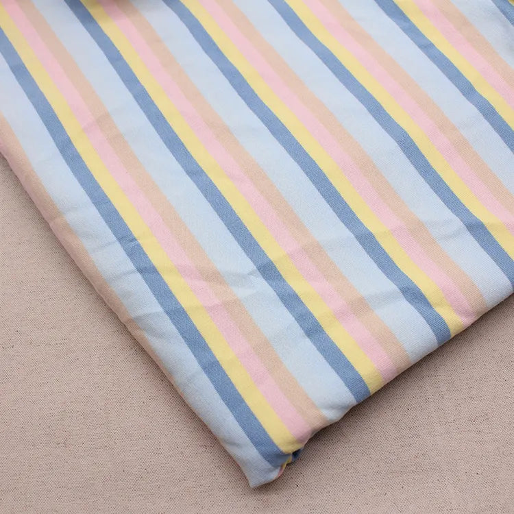 Un morceau plié de Longan Craft France Tissu French Terry extensible à rayures arc-en-ciel, présentant des rayures verticales bleues, jaunes, roses et beiges, est présenté sur une surface beige unie.