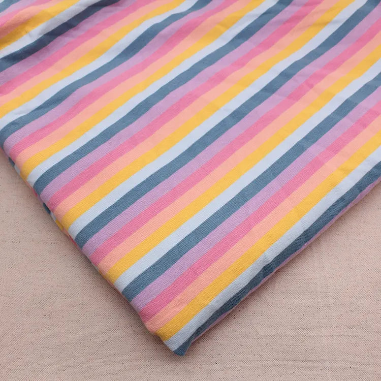 Un morceau de Longan Craft France Tissu French Terry extensible à rayures arc-en-ciel (coton tricoté, doux) est plié sur une surface beige, mettant en valeur ses rayures diagonales roses, jaunes, blanches et bleues.