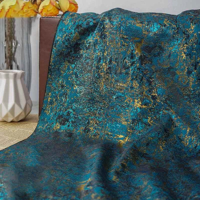 Un morceau de Tissu brocart vert rétro au mètre de Longan Craft France, avec des détails métalliques en jacquard doré, est drapé sur une surface brune ; à l'arrière-plan se trouvent un vase géométrique blanc et des fleurs jaunes en verre.