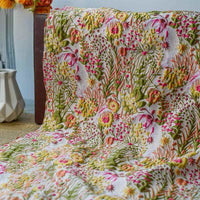 Un jeté en tissu brocart rétro floral au mètre de Longan Craft France, avec des motifs jacquard verts, jaunes et roses, est drapé sur du bois. Un vase blanc ajoute un charme vintage à l'arrière-plan gauche.