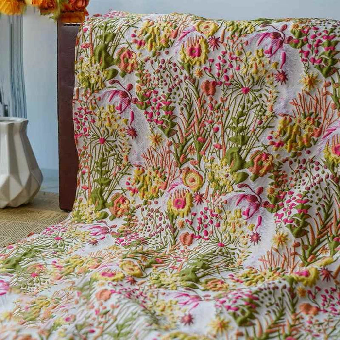 Un jeté en tissu brocart rétro floral au mètre de Longan Craft France, avec des motifs jacquard verts, jaunes et roses, est drapé sur du bois. Un vase blanc ajoute un charme vintage à l'arrière-plan gauche.