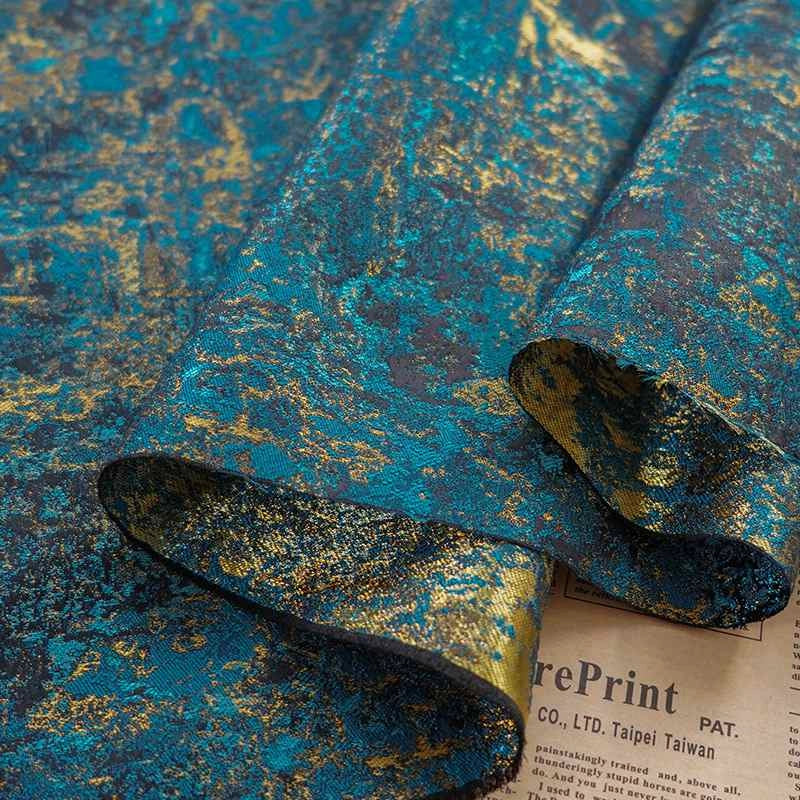 Gros plan sur le Tissu brocart vert rétro au mètre de Longan Craft France, un jacquard d'ameublement aux accents métalliques dorés et au motif marbré, drapé sur un journal.