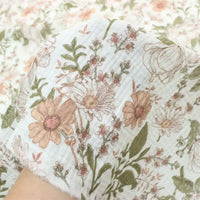 Une main tient un morceau de Double gaze à fleurs rétro au mètre de Longan Craft France, un tissu floral de style vintage 100 % coton, dans les tons rose, beige et vert, avec d'autres pièces de ce charmant tissu à l'arrière-plan.