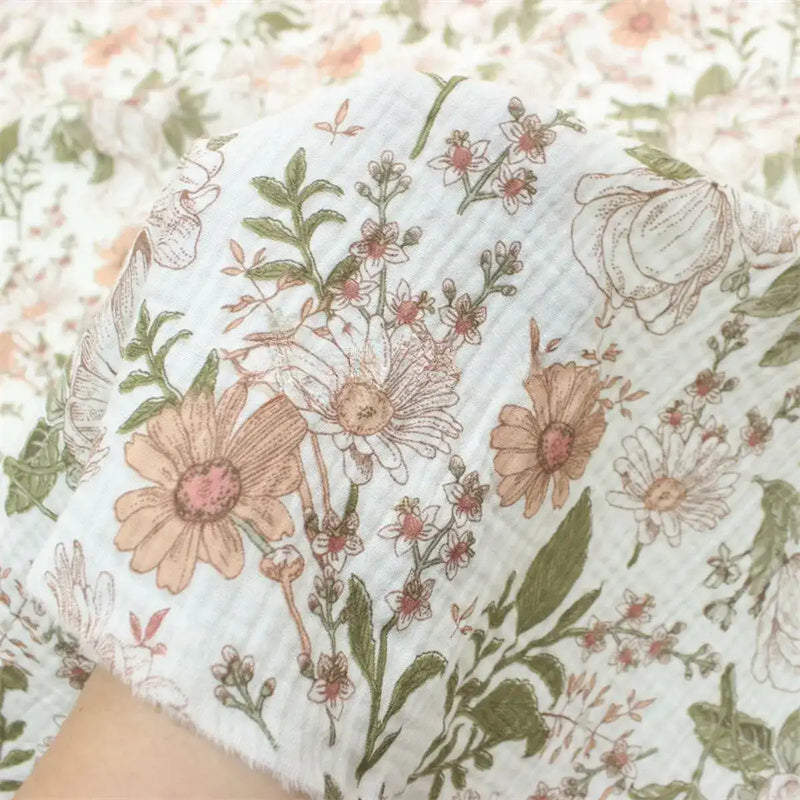 Une main tient un morceau de Double gaze à fleurs rétro au mètre de Longan Craft France, un tissu floral de style vintage 100 % coton, dans les tons rose, beige et vert, avec d'autres pièces de ce charmant tissu à l'arrière-plan.
