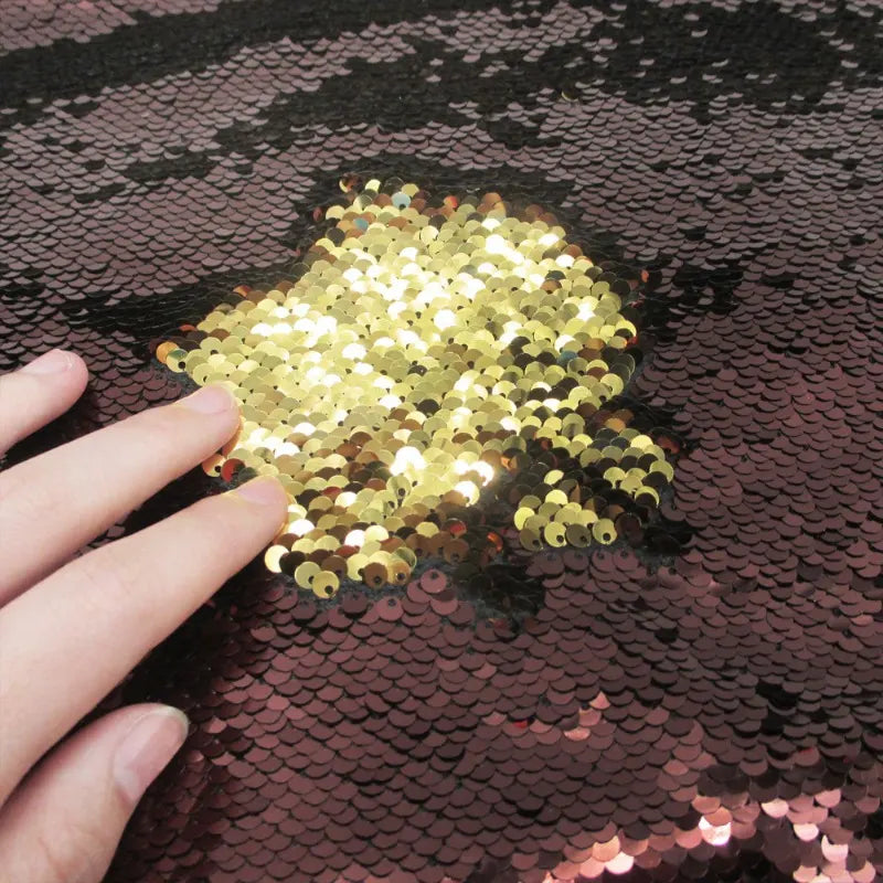 Une main effleure le Tissu pailleté bicolore réversible au mètre de Longan Craft France, révélant des paillettes dorées étincelantes sous une couche de paillettes réversibles rose foncé ou marron.