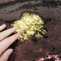 Une main effleure le Tissu pailleté bicolore réversible au mètre de Longan Craft France, révélant des paillettes dorées étincelantes sous une couche de paillettes réversibles rose foncé ou marron.