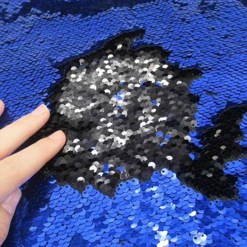 Une main effleure le Tissu pailleté bicolore réversible au mètre de Longan Craft France, révélant des paillettes argentées et noires sous des paillettes bleues sur ce tissu à paillettes réversibles scintillant, parfait pour éblouir les accessoires.