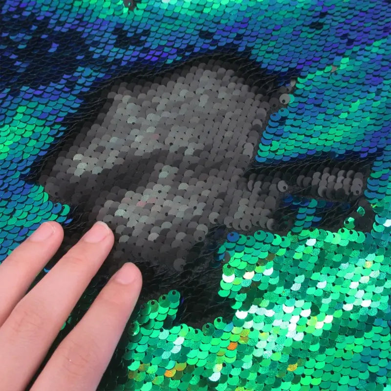 Une main passe sur le tissu pailleté bicolore réversible au mètre de Longan Craft France, faisant passer les paillettes du vert et du bleu vifs au noir, révélant une tache sombre au milieu de la surface brillante et étincelante.