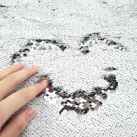Une main dessine un cœur sur le tissu pailleté bicolore réversible au mètre de Longan Craft France, révélant des paillettes plus sombres sous des paillettes argentées brillantes.