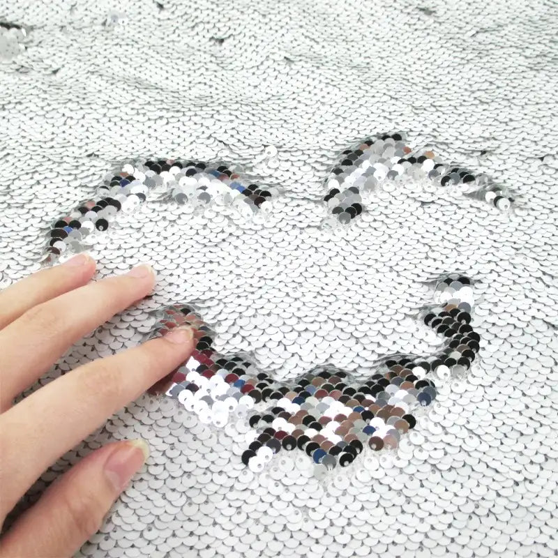 Une main dessine un cœur sur le tissu pailleté bicolore réversible au mètre de Longan Craft France, révélant des paillettes plus sombres sous des paillettes argentées brillantes.
