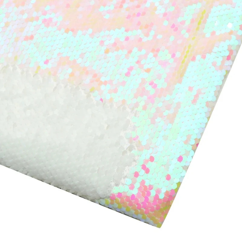 Un gros plan montre le tissu pailleté bicolore réversible au mètre de Longan Craft France, dont un coin est retourné pour révéler le dessous. Des paillettes blanches irisées reflètent des teintes pastel roses, jaunes et vertes.