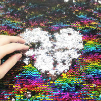 Une main effleure le Tissu pailleté bicolore réversible au mètre de Longan Craft France, révélant un cœur blanc parmi les paillettes multicolores de ce tissu réversible scintillant pour accessoires.