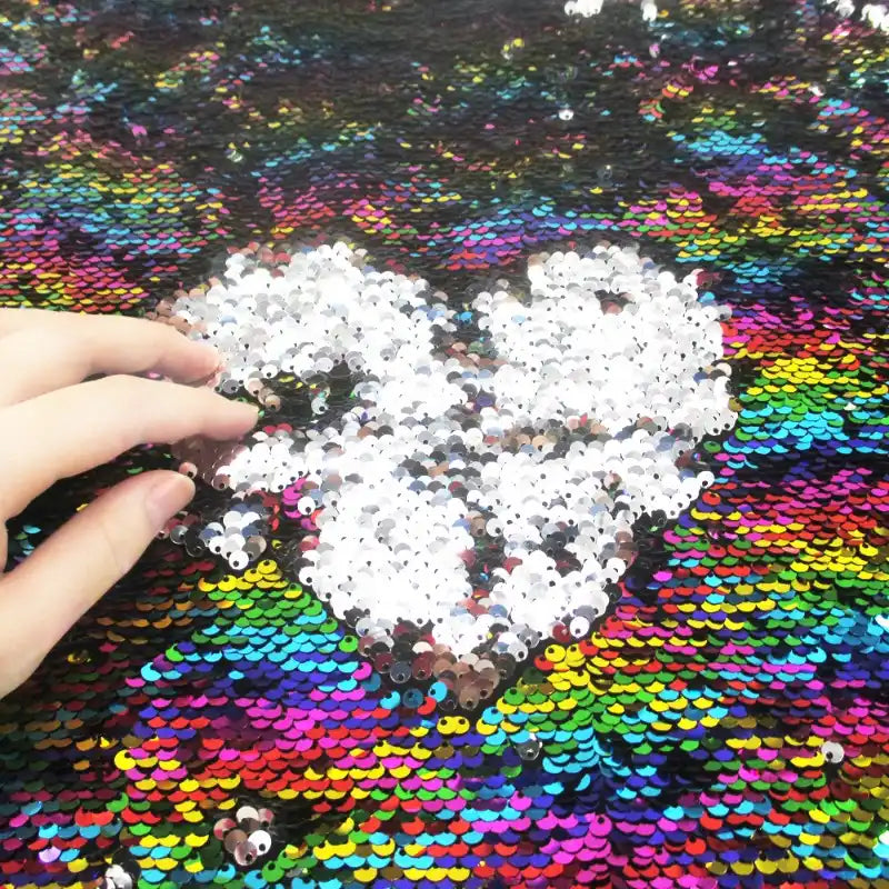 Une main effleure le Tissu pailleté bicolore réversible au mètre de Longan Craft France, révélant un cœur blanc parmi les paillettes multicolores de ce tissu réversible scintillant pour accessoires.