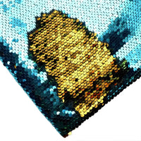 Gros plan sur un visage en paillettes d'or sur fond bleu et blanc, réalisé avec le Tissu pailleté bicolore réversible au mètre de Longan Craft France, un tissu à paillettes réversibles chatoyant, vu de biais.