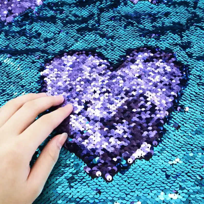 Une main effleure le tissu pailleté bicolore réversible de Longan Craft France, créant un cœur en paillettes violettes sur fond bleu - un tissu scintillant idéal pour les accessoires et les travaux manuels.
