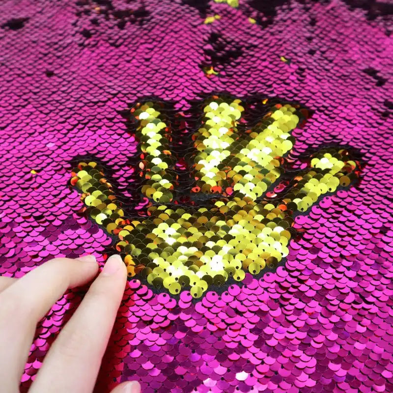 Une main effleure des paillettes sur le Tissu pailleté bicolore réversible au mètre de Longan Craft France, révélant une empreinte de main dorée sur un fond de paillettes roses.