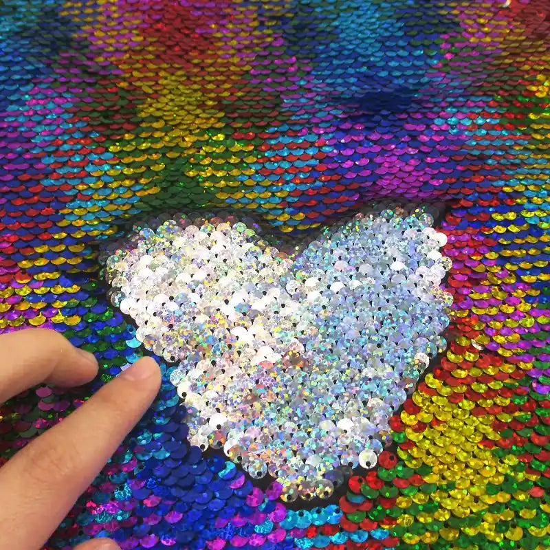 Une main passe les paillettes sur un Tissu pailleté bicolore réversible au mètre à motifs arc-en-ciel de Longan Craft France, révélant un cœur en paillettes blanches brillantes - parfait pour des accessoires étincelants.