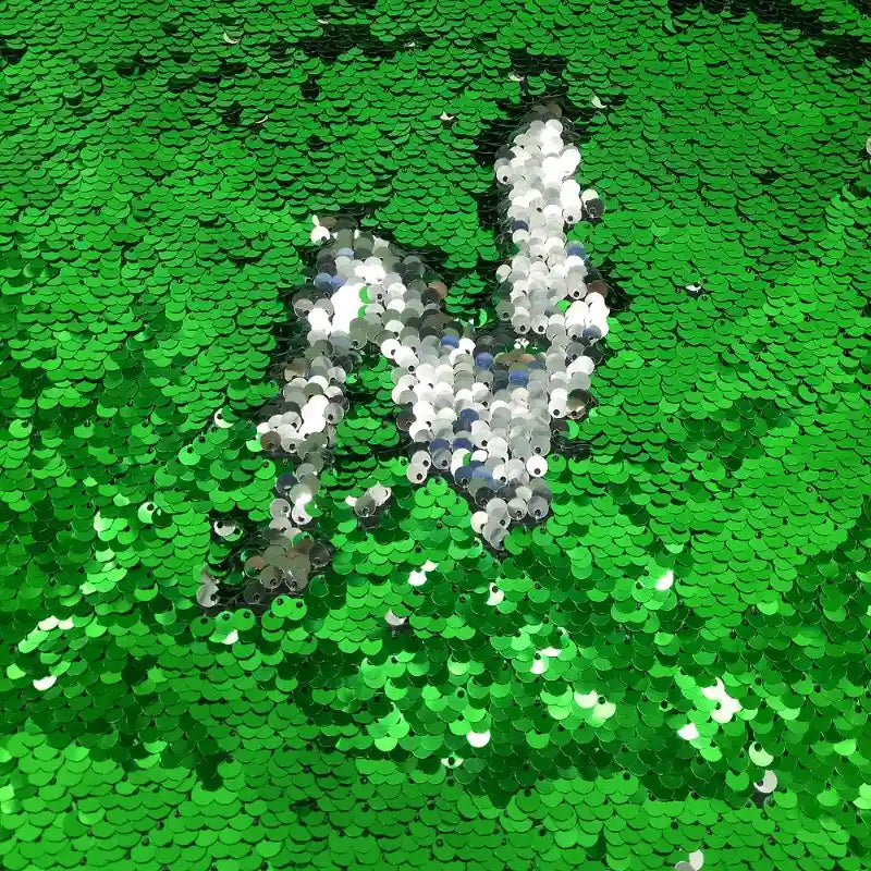 Un "Tissu pailleté bicolore réversible au mètre" vert de Longan Craft France, avec des paillettes brossées en argent en dessous, forme la lettre "N".