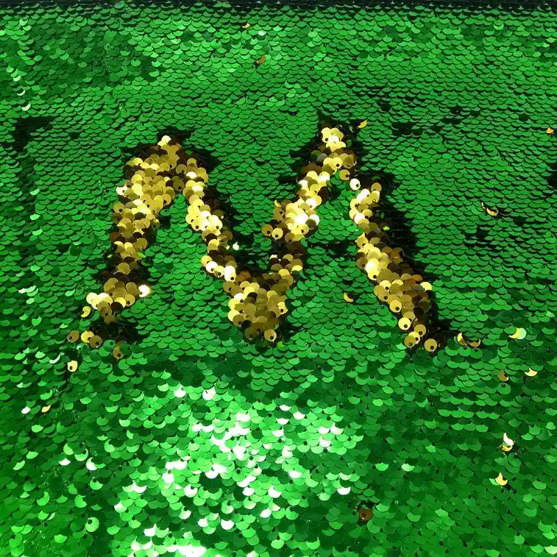Le Tissu pailleté bicolore réversible au mètre de Longan Craft France est orné de paillettes vertes et dorées, l'or formant un "M" réfléchissant au centre. Ce tissu éblouissant capte magnifiquement la lumière - parfait pour les accessoires.