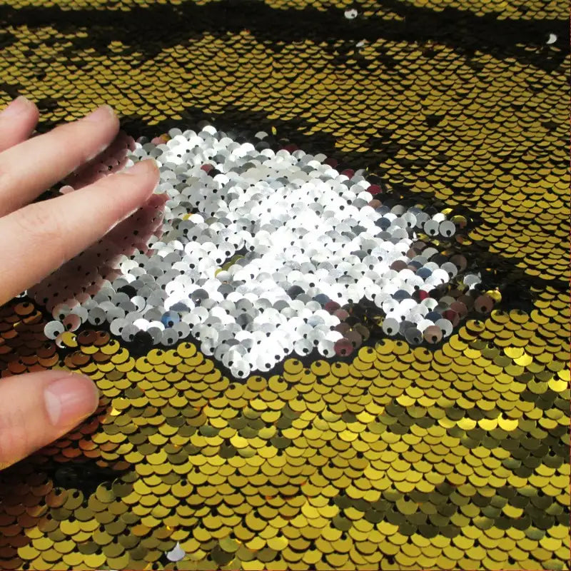 Une main passe sur le tissu pailleté bicolore réversible de Longan Craft France, transformant les paillettes majoritairement dorées pour révéler une tache d'argent étincelante parmi elles.