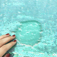 Une main avec du vernis à ongles rouge brosse le tissu pailleté bicolore réversible au mètre de Longan Craft France, révélant une partie mate et plus claire lorsque les paillettes se retournent pour montrer leur autre face.
