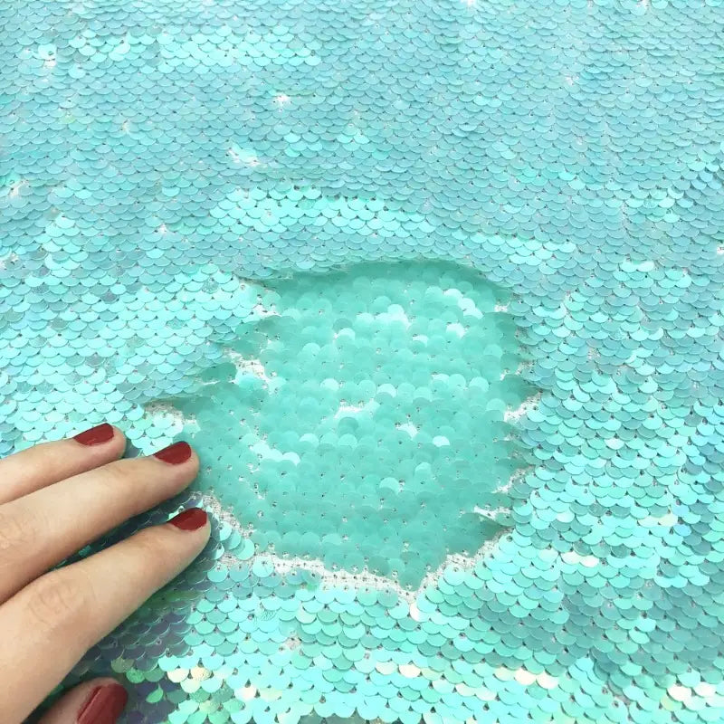 Une main avec du vernis à ongles rouge brosse le tissu pailleté bicolore réversible au mètre de Longan Craft France, révélant une partie mate et plus claire lorsque les paillettes se retournent pour montrer leur autre face.