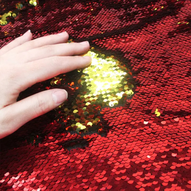 Une main glisse sur le tissu pailleté bicolore réversible de Longan Craft France, révélant une partie dorée parmi des paillettes rouges, une matière éblouissante et réversible parfaite pour créer des accessoires étincelants.